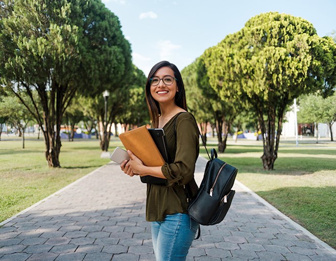 Smiling female college student wearing backpack and holding laptop and phone outside