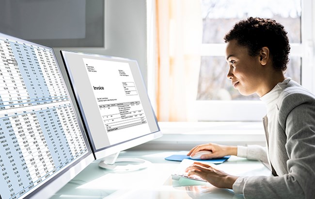 Woman sitting at desk looking at two large computer monitors looking at spreadsheets