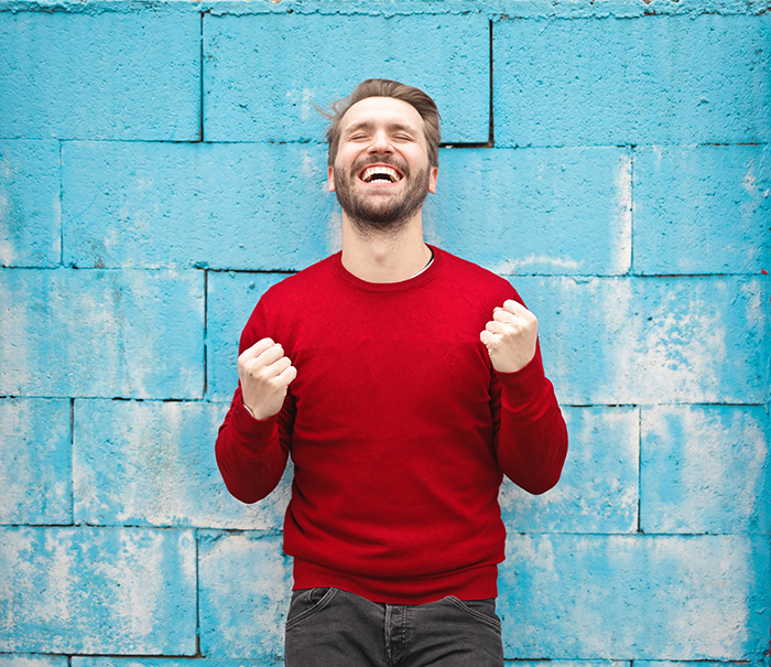 Man with eyes closed and fists clenched smiling in front of blue brick building