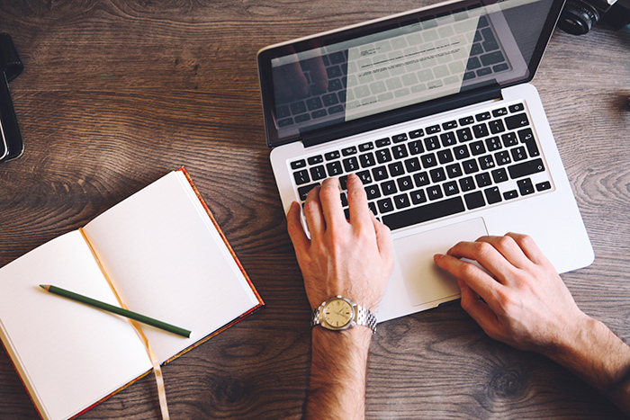 Two hands typing on laptop alongside an empty notebook with pencil on top