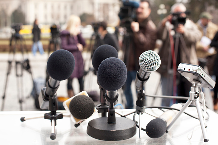 Multiple microphones on table with reporters blurred in the background with cameras