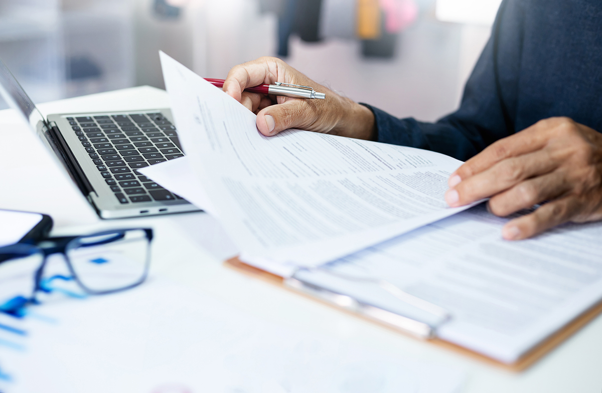 Close up of a desk with a stack of papers in a clipboard and an open laptop
