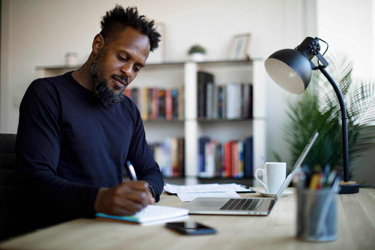Man sitting writing on paper at a desk with an open laptop