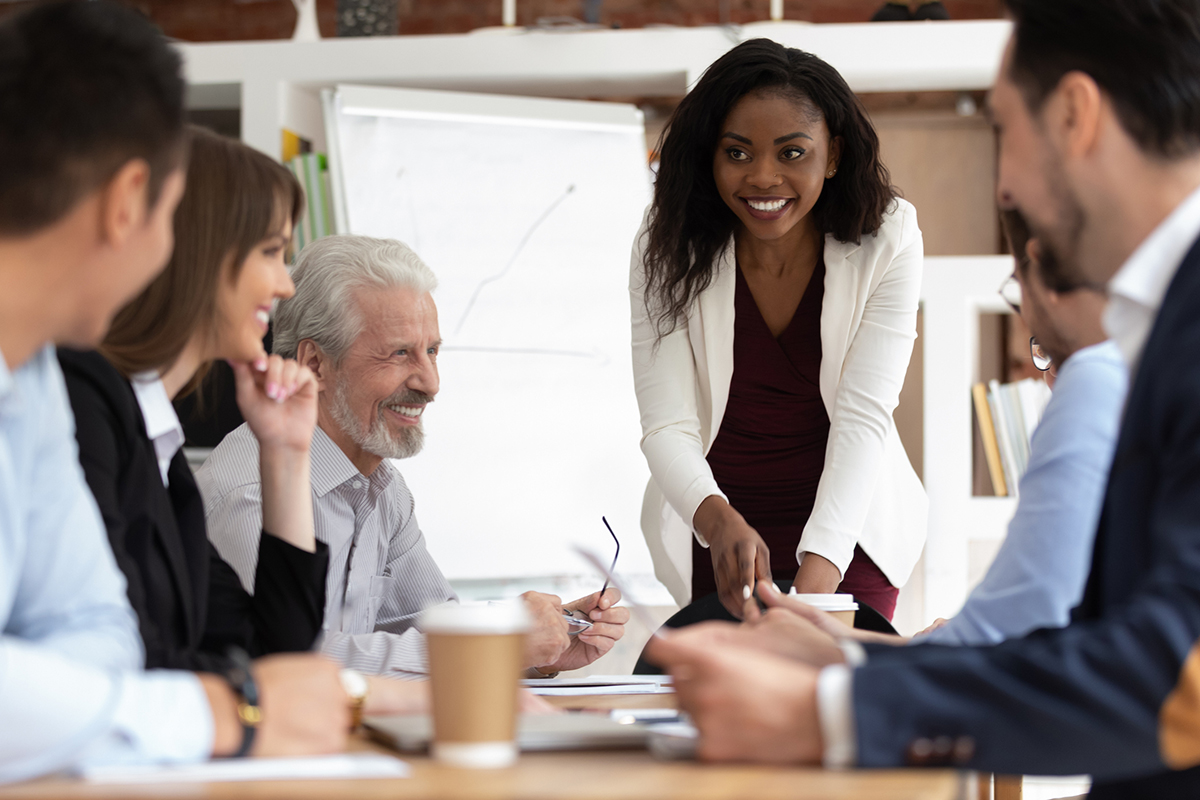 smiling woman at front of conference table giving presentation to coworkers