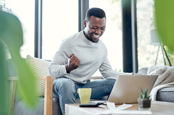 Excited man celebrating with clenched fist sitting on couch with open laptop on coffee table 