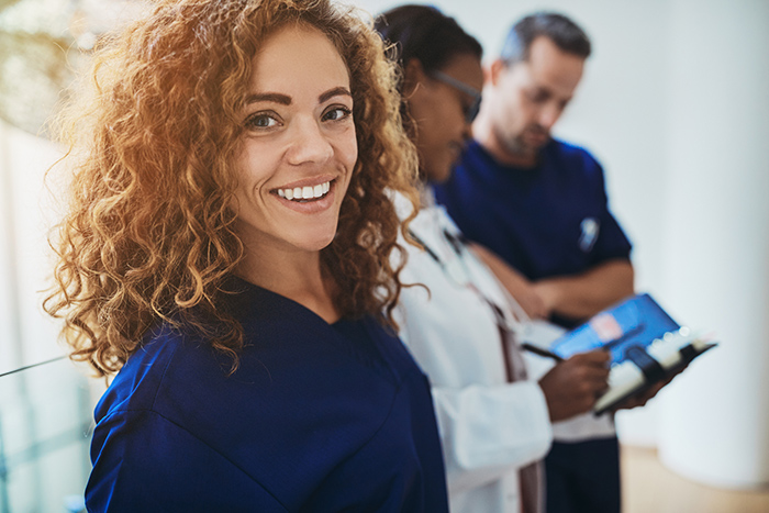 Smiling woman in navy blue scrubs looking at camera with two healthcare workers in background
