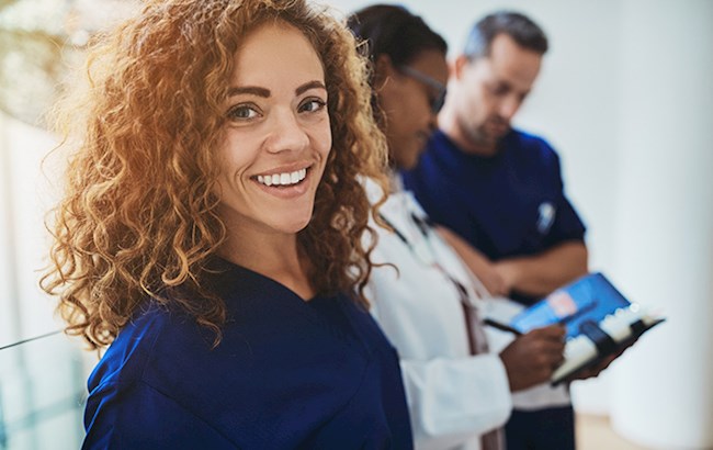 Smiling woman in navy blue scrubs looking at camera with two healthcare workers in background