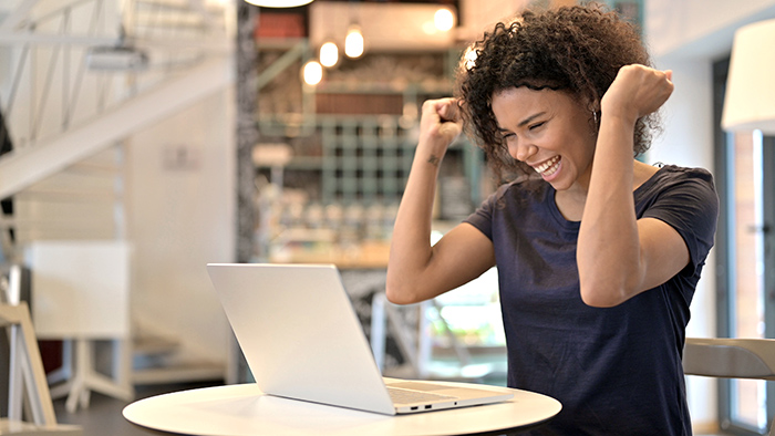 Woman celebrating with two clenched fists in the air looking at open laptop screen inside office