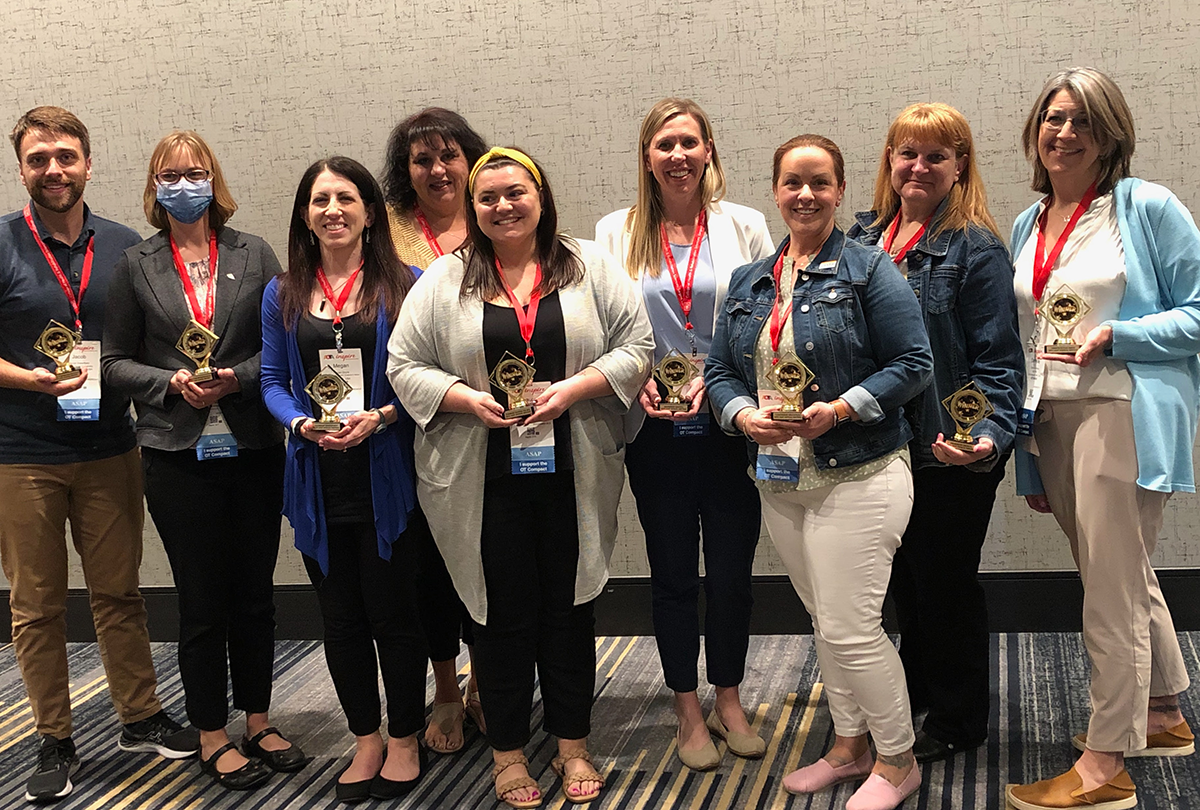 Nine people wearing AOTA INSPIRE badges with orange lanyards and holding small gold trophies. The trophies have the word "winner" written on the front.