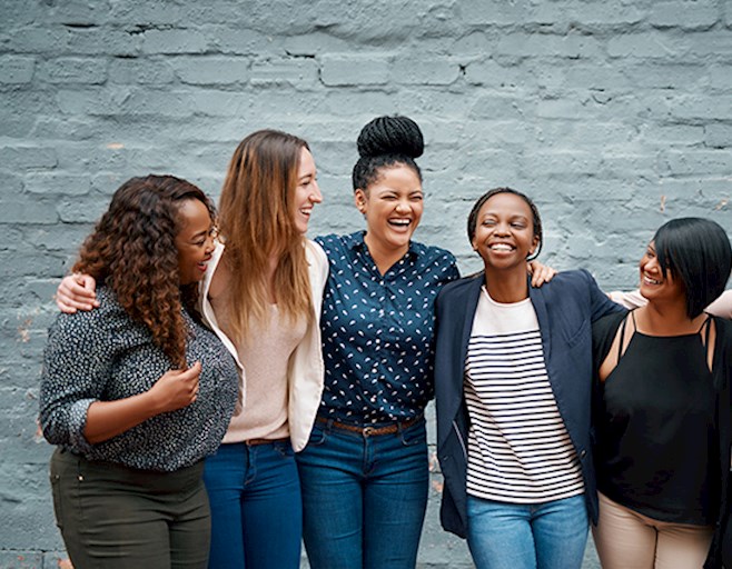 Diverse group of woman with arms around each other smiling in front of gray brick wall