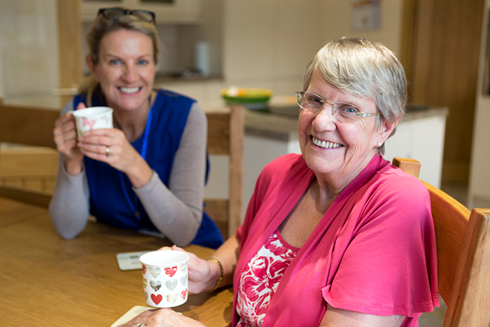 Female healthcare professional sitting at table drinking tea with senior woman smiling at camera