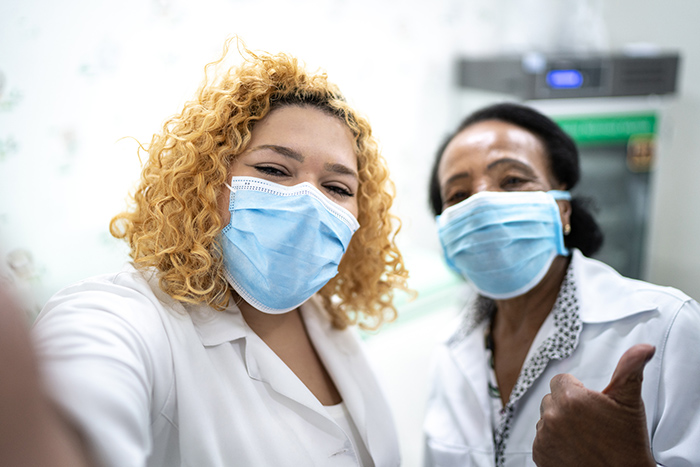 Two healthcare workers wearing face masks taking selfie and giving thumbs up