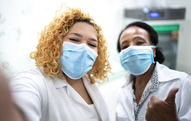 Two healthcare workers wearing face masks taking selfie and giving thumbs up
