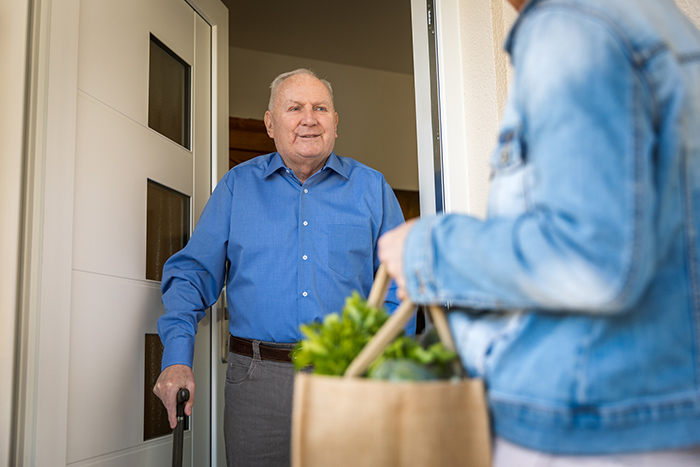 Senior man using cane standing in doorway greeting woman holding bag of food
