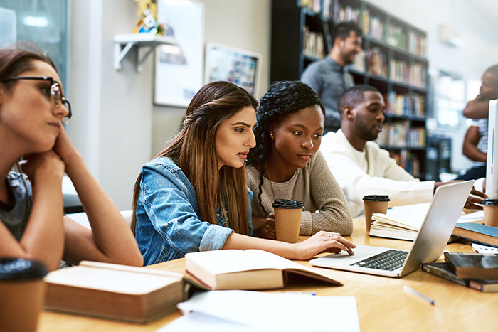 Two women studying at laptop inside library with others in the background