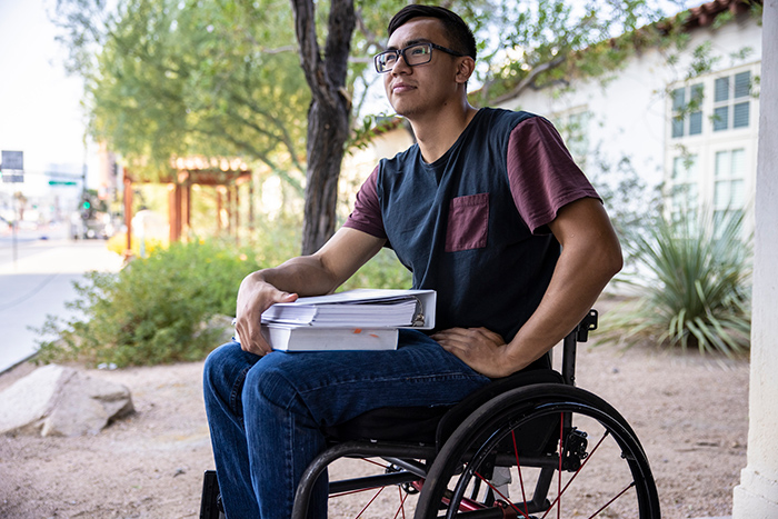 Male college student outside in a wheelchair holding a binder and a textbook