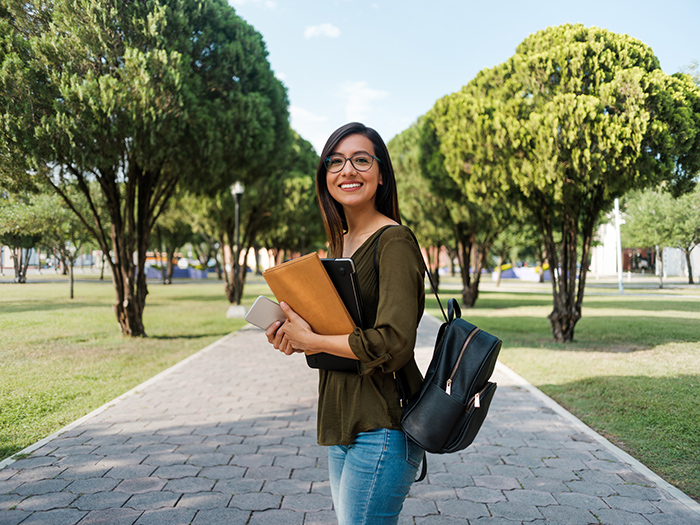 Smiling female college student wearing backpack and holding laptop and phone outside