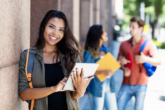 Female college student holding tablet smiling at camera with two friends talking in background outside