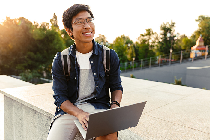 Male college student sitting outside wearing backpack while using laptop