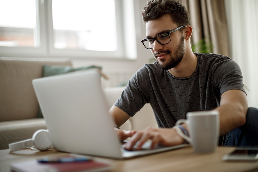Smiling man wearing glasses sitting at desk working on open laptop 