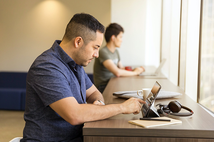 Man working on tablet at high table in front of a window with someone on laptop in background