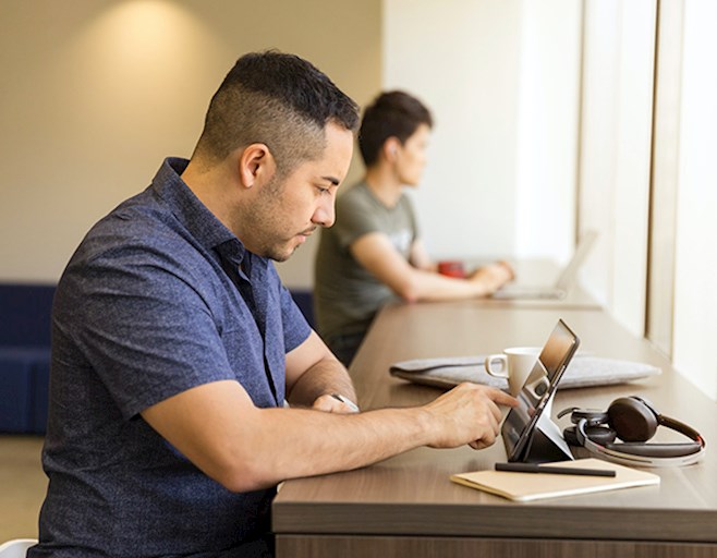 Man working on tablet at high table in front of a window with someone on laptop in background