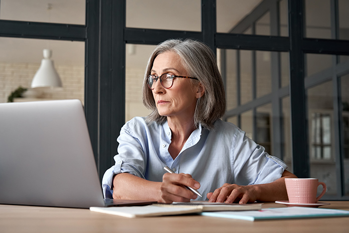 Older woman doing paperwork at table with open laptop and cup of coffee