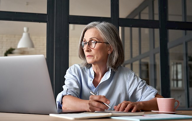 Older woman doing paperwork at table with open laptop and cup of coffee