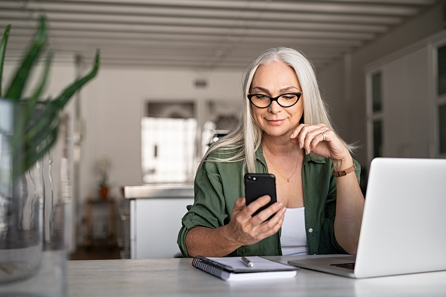 Mature woman with long gray hair sitting at desk working on laptop and reading her phone