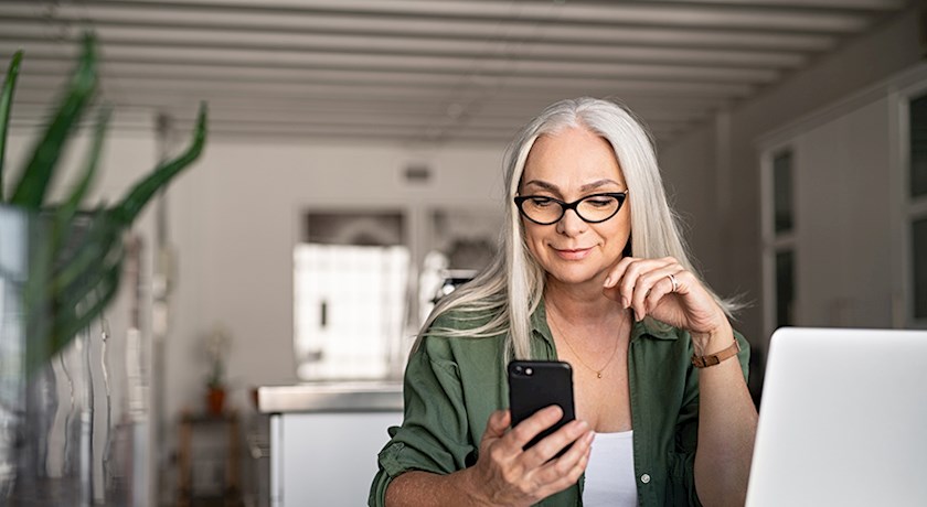 Mature woman with long gray hair sitting at desk working on laptop and reading her phone
