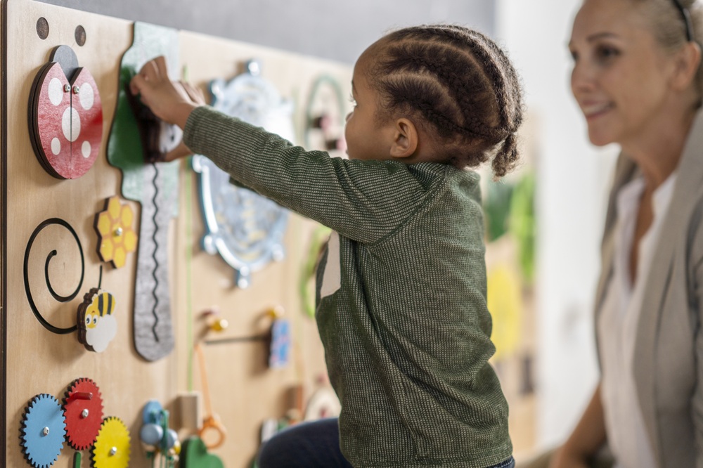 Preschool-aged child plays with sensory board during occupational therapy session while occupational therapy practitioner looks on.