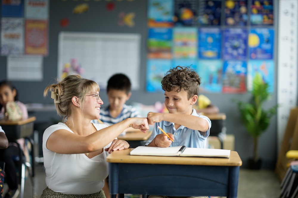 Practitioner in a school classroom setting crouched down next to desk with young child. The two are fist bumping in the air over the desk.