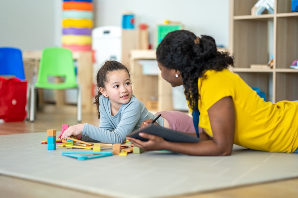 Occupational therapy practitioner and young child both lying on their stomachs on a mat on the floor while the child plays with blocks.