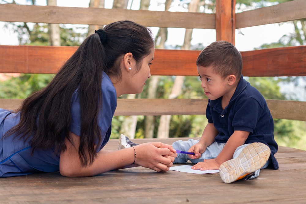 Occupational therapy practitioner lays on her stomach while young child sits across from her coloring in a notebook. They are sitting outside on a deck.