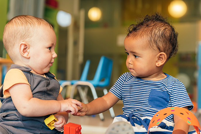 Two babies sitting up holding hands and playing with toys