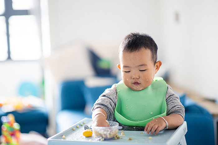 Baby in high chair with food on face and hand in bowl eating