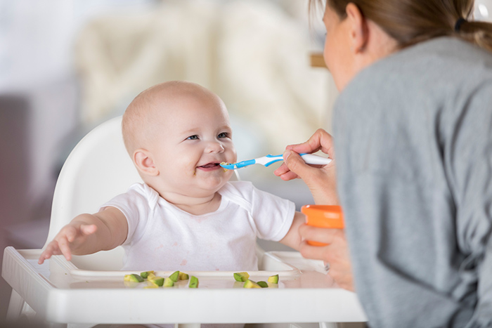 Baby in high chair smiling as woman feeds him with blue spoon