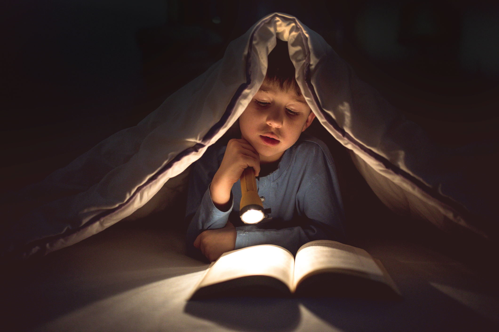 Child reading a book in bed under the covers with a flashlight
