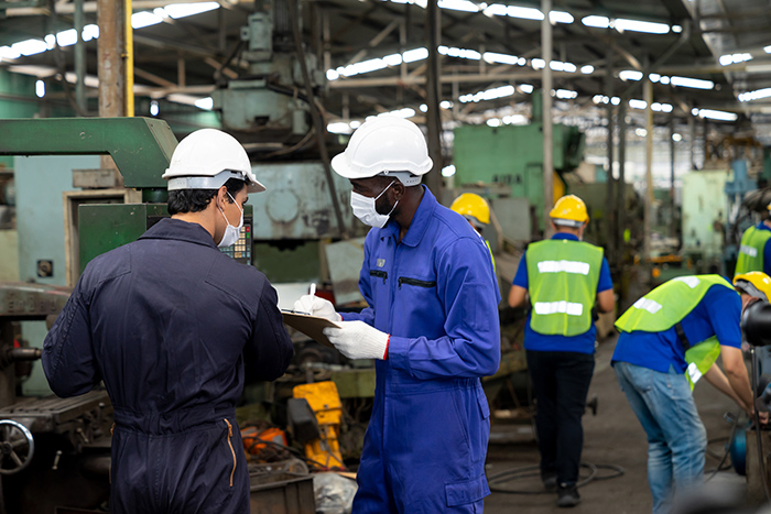 Two men wearing face masks and jumpsuits working inside a factory with others working in the background