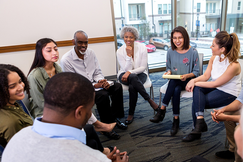 Diverse group of people sitting in a circle talking in chairs inside a room