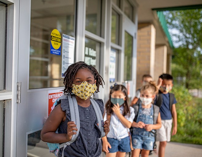 Young children wearing face masks wearing backpacks lined up outside of school building