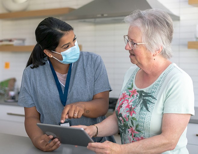 Female occupational therapist wearing a face mask helping senior client in kitchen reading materials on a tablet