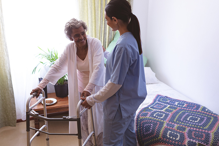 Female occupational therapist helping senior woman get out of bed and use walker