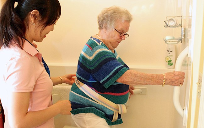 Senior woman stepping into adaptive shower holding grip bar with occupational therapist holding support belt