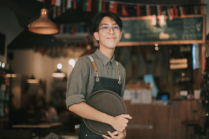 Smiling teenage boy wearing apron holding a round tray inside a restaurant