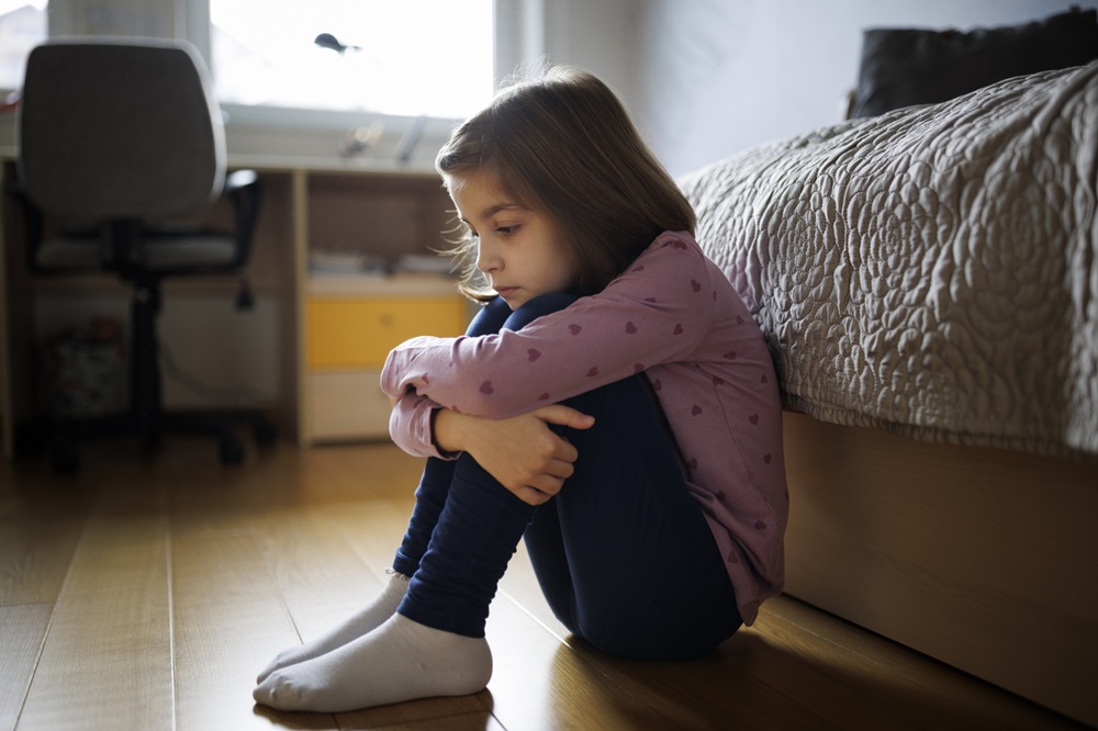 Child sitting on the floor at the foot of a bed with legs up against chest 