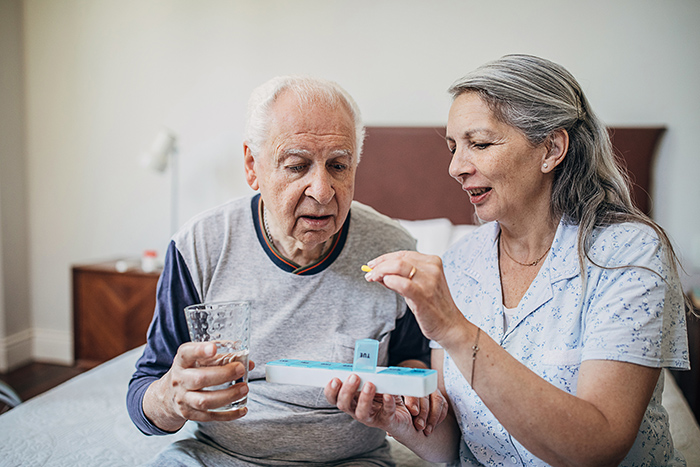 Woman holding pill organizer helping man beside her holding water take medication
