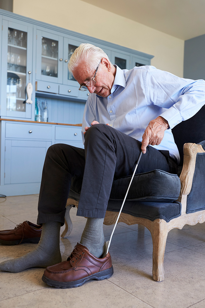Older man sitting in chair putting on shoe while using long gripper