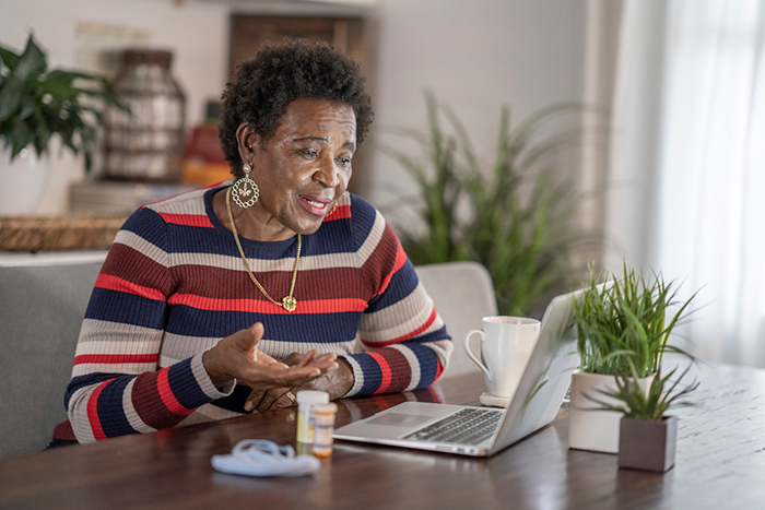Senior woman at table talking into laptop with medication beside her