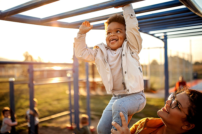 Woman holding up smiling toddler to monkey bars outside at park